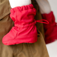 A close-up of a bright red baby boot with a drawstring, shown hanging from the waist of a person wearing tan pants and white socks.