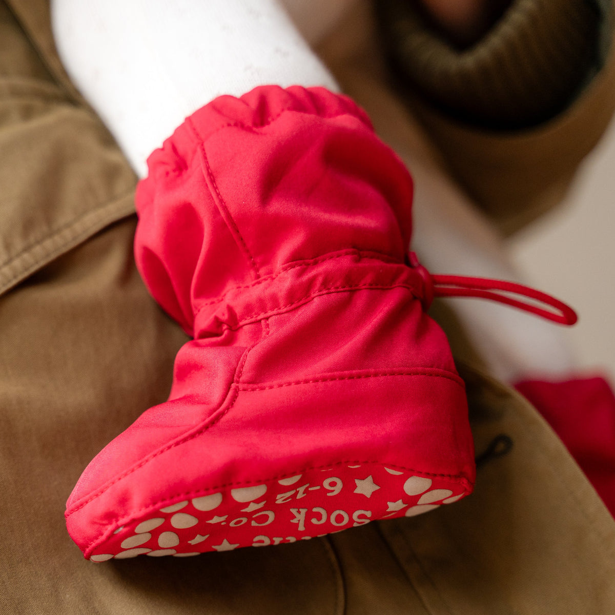A close-up of a bright red knit bootie with a drawstring and a white patterned sole, shown resting on tan pants.