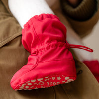 A close-up of a bright red knit bootie with a drawstring and a white patterned sole, shown resting on tan pants.