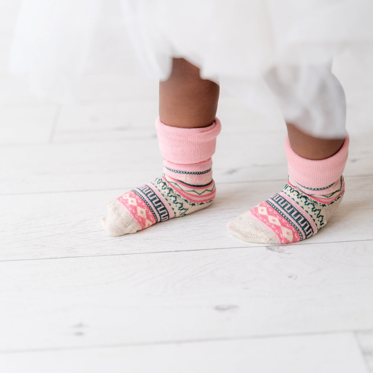 A close-up of a child's feet wearing pink and multicolor patterned socks with a white dress hem visible above them, standing on a white wooden floor.