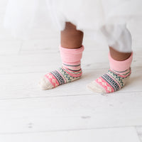 A close-up of a child's feet wearing pink and multicolor patterned socks with a white dress hem visible above them, standing on a white wooden floor.