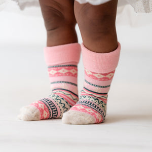 A close-up of a child's legs wearing pink knit socks with a multicolor geometric pattern, standing on a white floor against a white background.