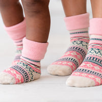 A close-up of a child's legs wearing pink patterned socks with geometric Fair Isle designs in pink, navy, and cream, standing on a light wooden floor.